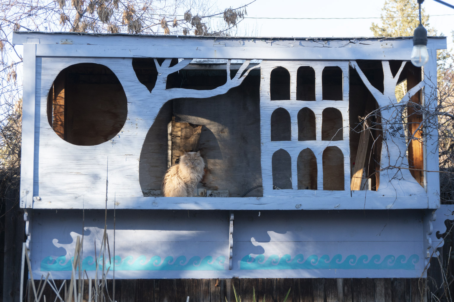 A cat sits in a blue wood structure with cutouts of trees and windows